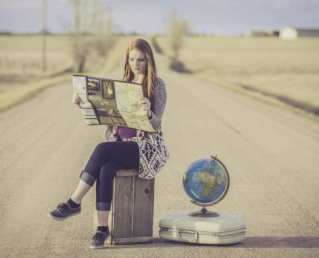 En ung kvinde sidder på en kasse midt på en landevej og kigger på kort. Ved siden af hende står en kuffert og en globus.