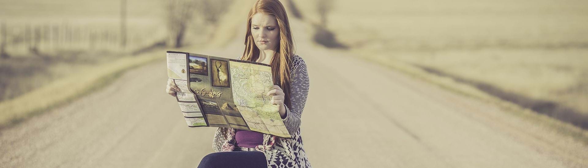En ung kvinde sidder på en kasse midt på en landevej og kigger på kort. Ved siden af hende står en kuffert og en globus.