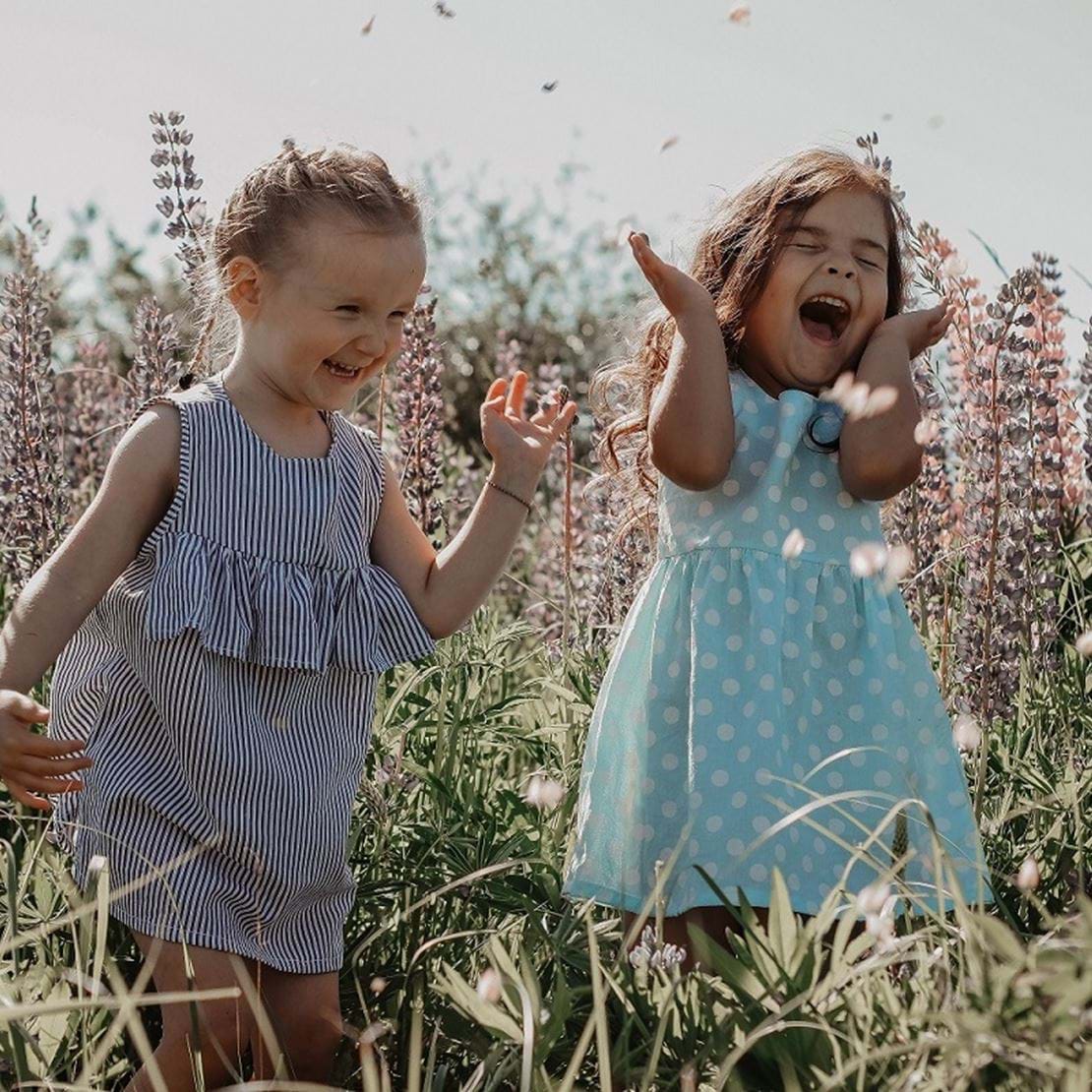 To små piger står på en blomstereng og griner hjerteligt og ser ud til at hygge sig meget.