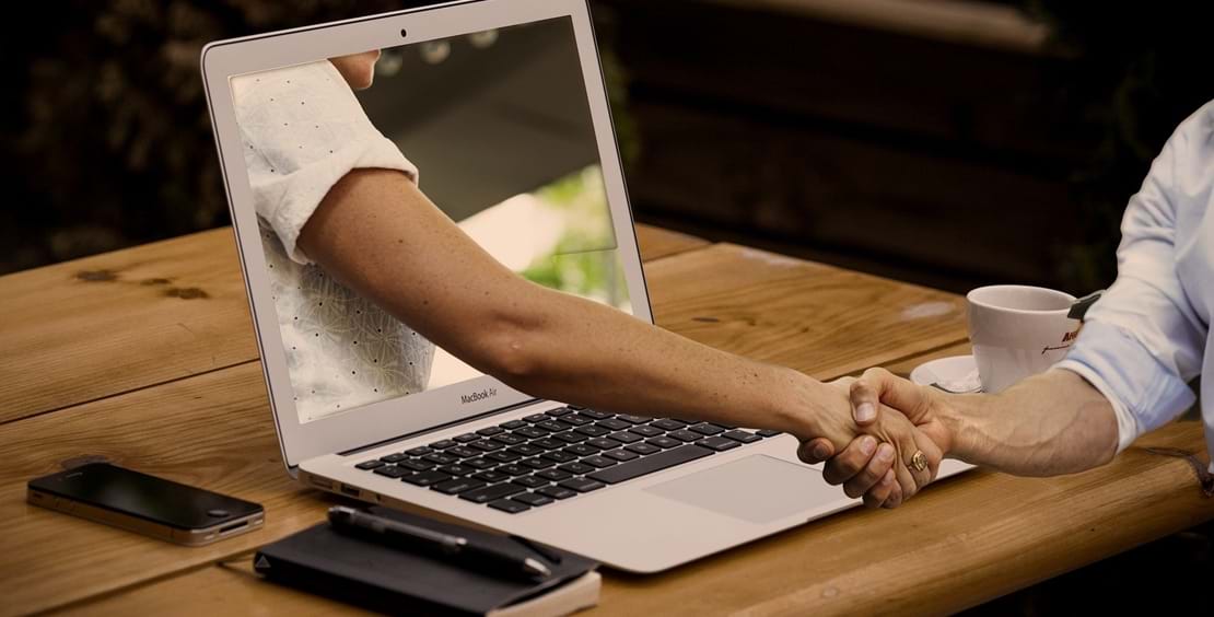 Et håndtryk i den virtuelle verden symboliseret ved en arm der kommer ud af skærmen på en laptop og trykker hånden på den person der sidder ved skærmen.