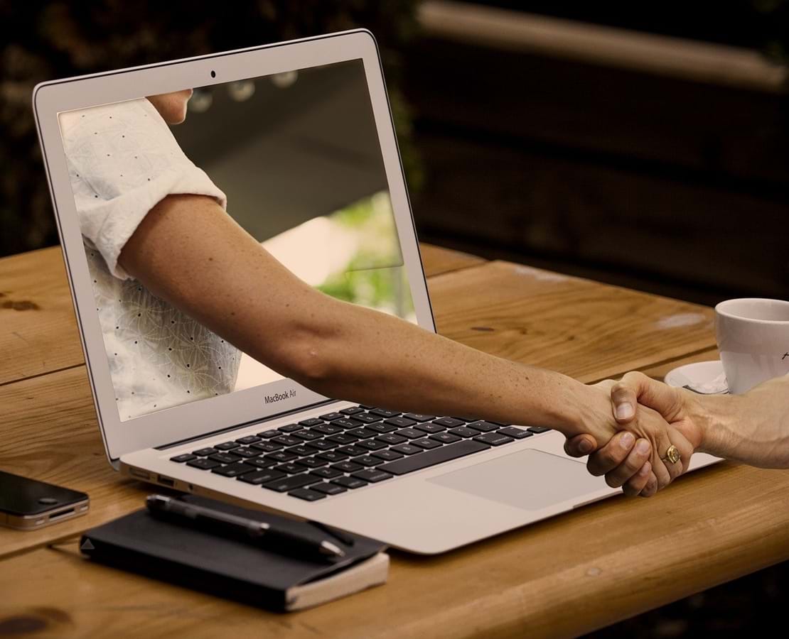 Et håndtryk i den virtuelle verden symboliseret ved en arm der kommer ud af skærmen på en laptop og trykker hånden på den person der sidder ved skærmen.