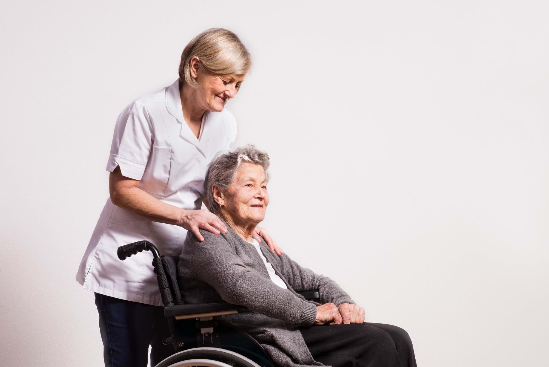 En kvinde i kittel skubber en ældre dame i kørestol.