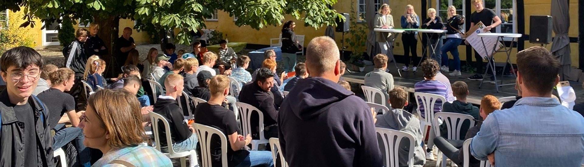 En forsamling af mennesker står og sidder med front op mod en scene, hvor der foregår en debat.