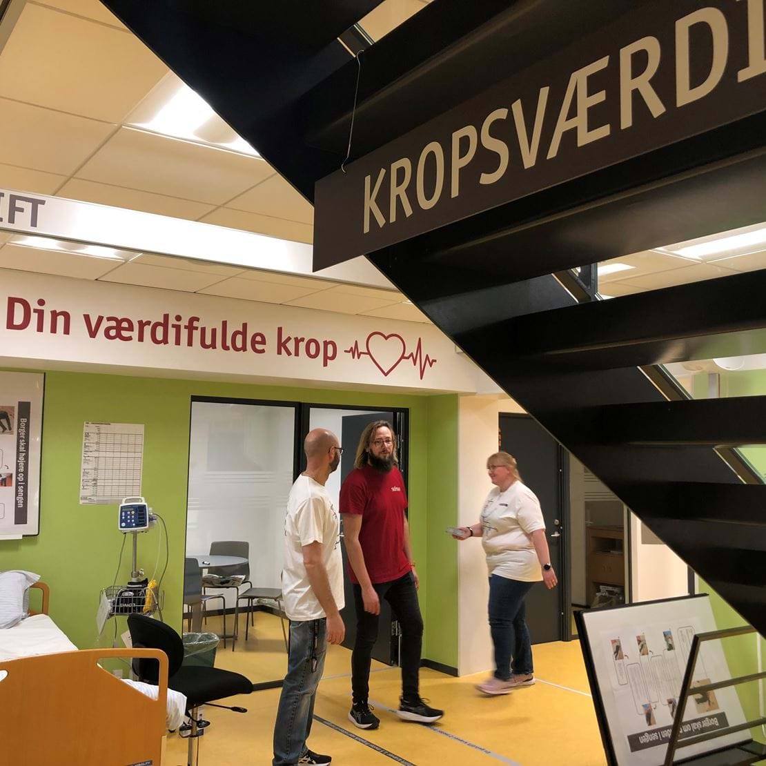 Her ses en af de første læringsstationer på SOSU Nykøbing Falster, hvor du kan styrke dine STEM-kompetencer og træne forflytning samt måling af kropsværdier. Tre medarbejdere figurerer på billedet sammen med en hospitalsseng og stationsskiltet "KROPSVÆRDI".