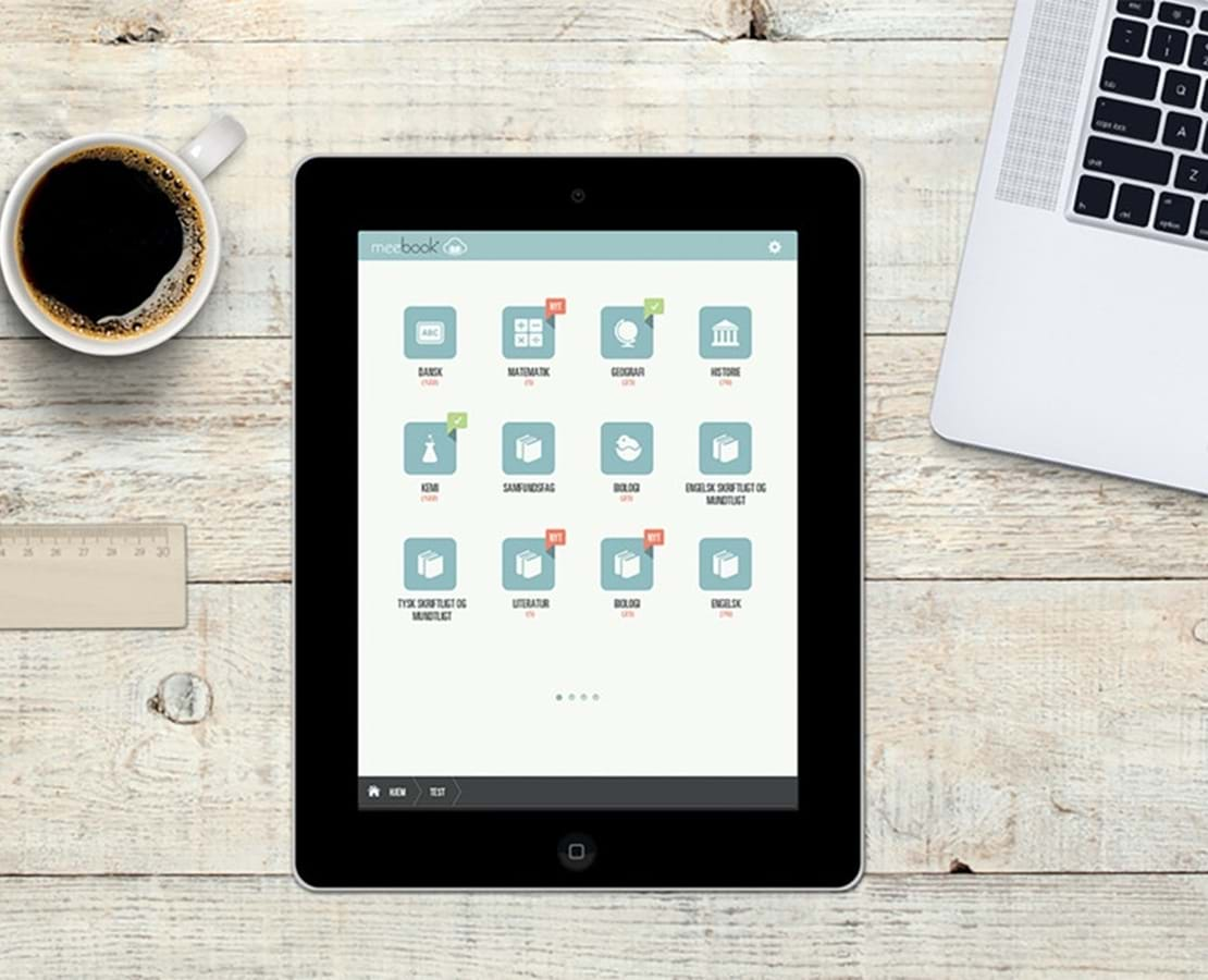 Skrivebord hvor en tablet med Meebook-app'en ligger i midten, en kop kaffe en laptop og et hæfte ligger uden om.