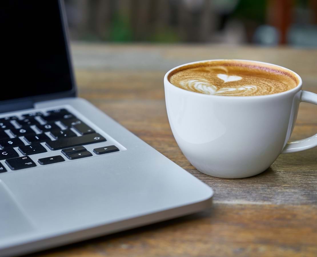 En kop cappucino med hjerte i mælkeskummet og en laptop ved siden af.