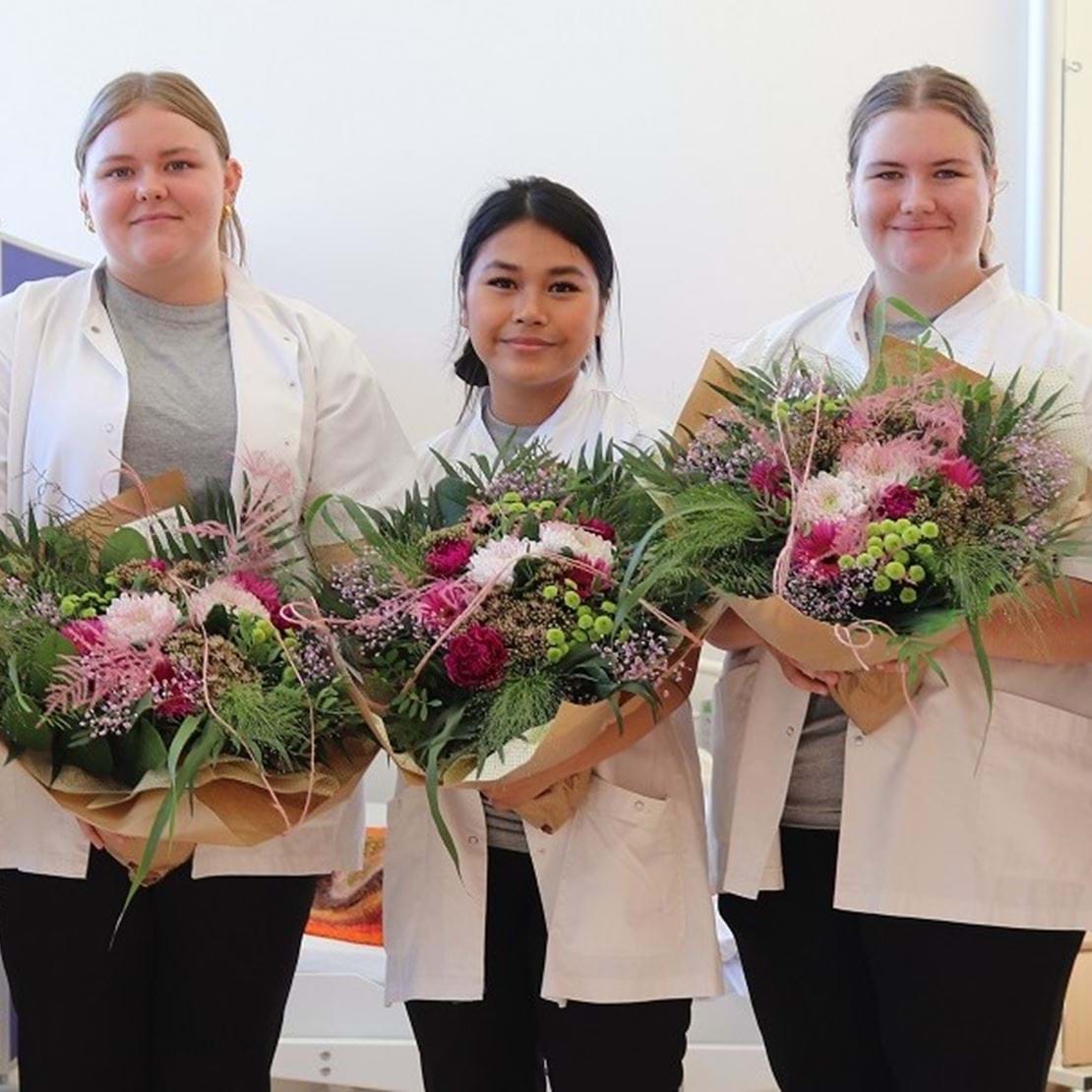 Fem glade piger står på rad og række med hver en blomsterbuket i hånden og kigger smilende ind i kameraet.