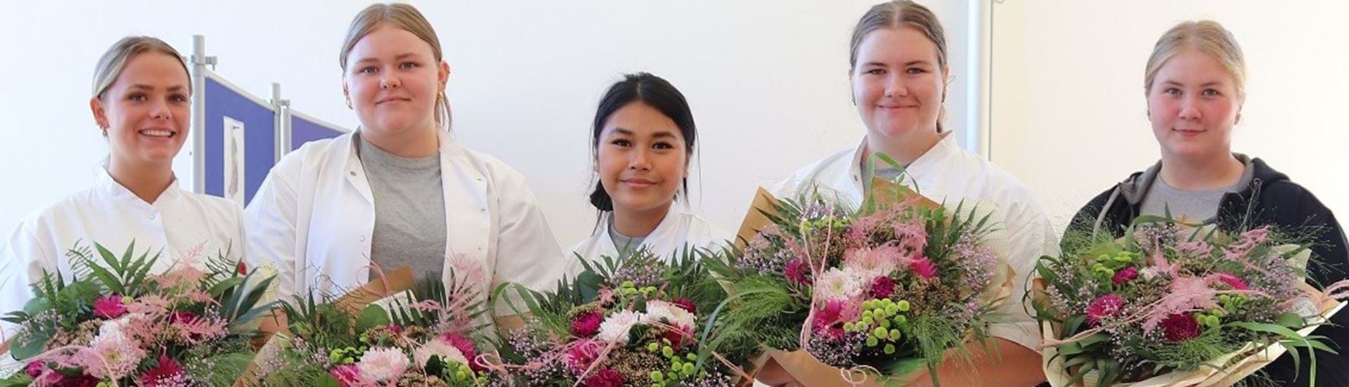 Fem glade piger står på rad og række med hver en blomsterbuket i hånden og kigger smilende ind i kameraet.