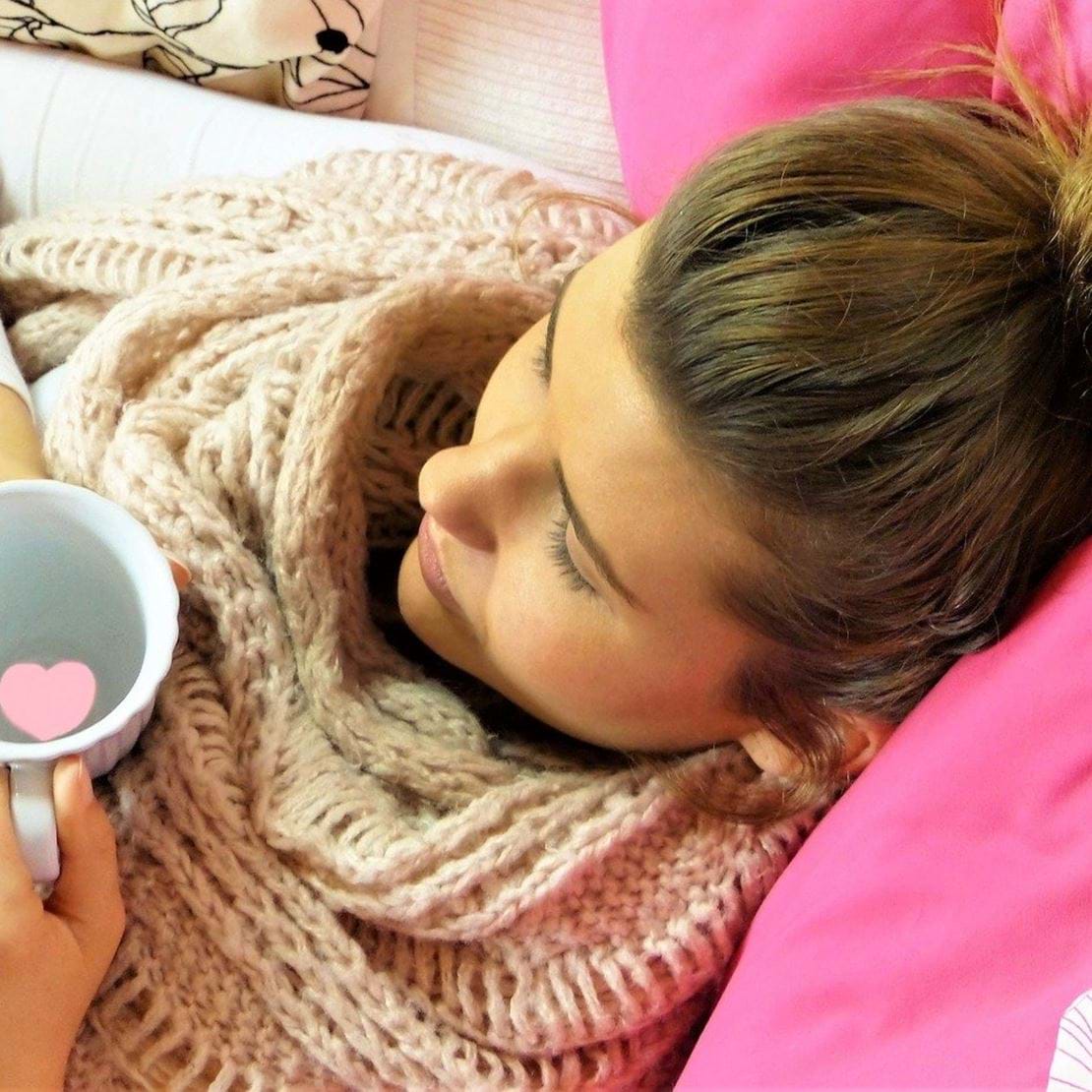 Pige ligger i sengen med halstørklæde på og en varm kop te med et lyserødt papirhjerte i