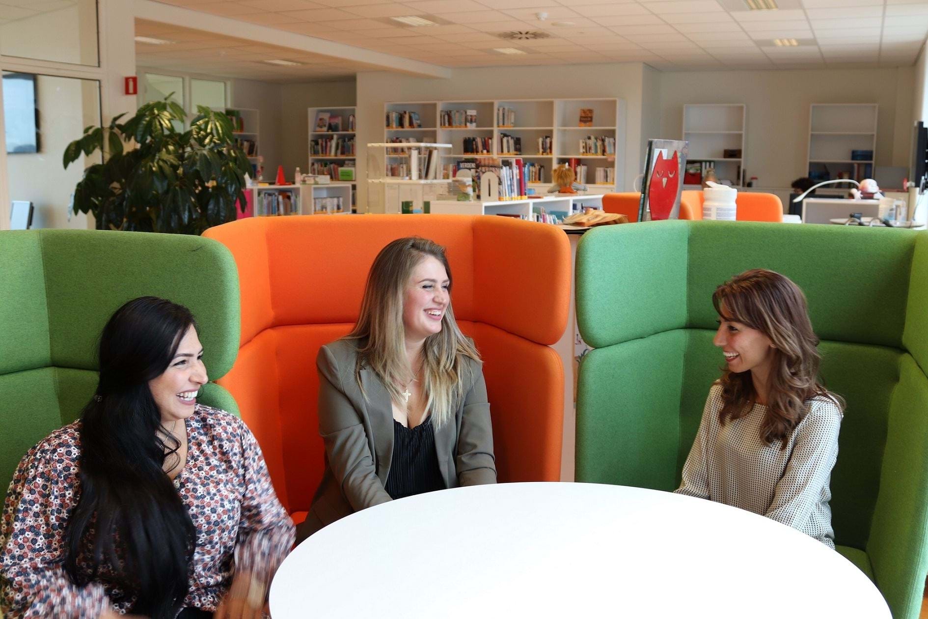 Tre voksne kvinder sidder i hver sin lænestol på et bibliotek og griner sammen.