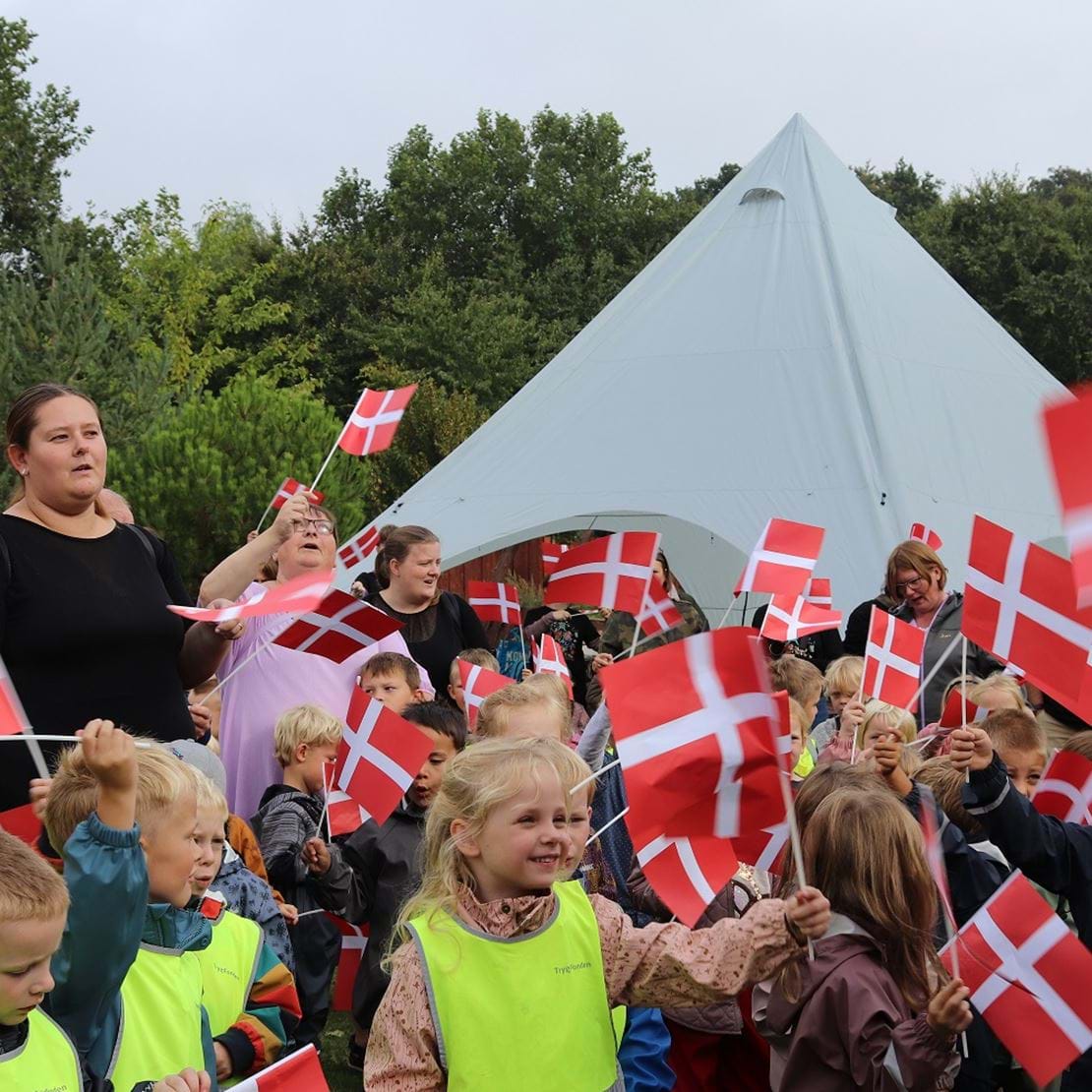 En stor mængde børn og voksne står med flag i hånden og synger fødselsdagssang.