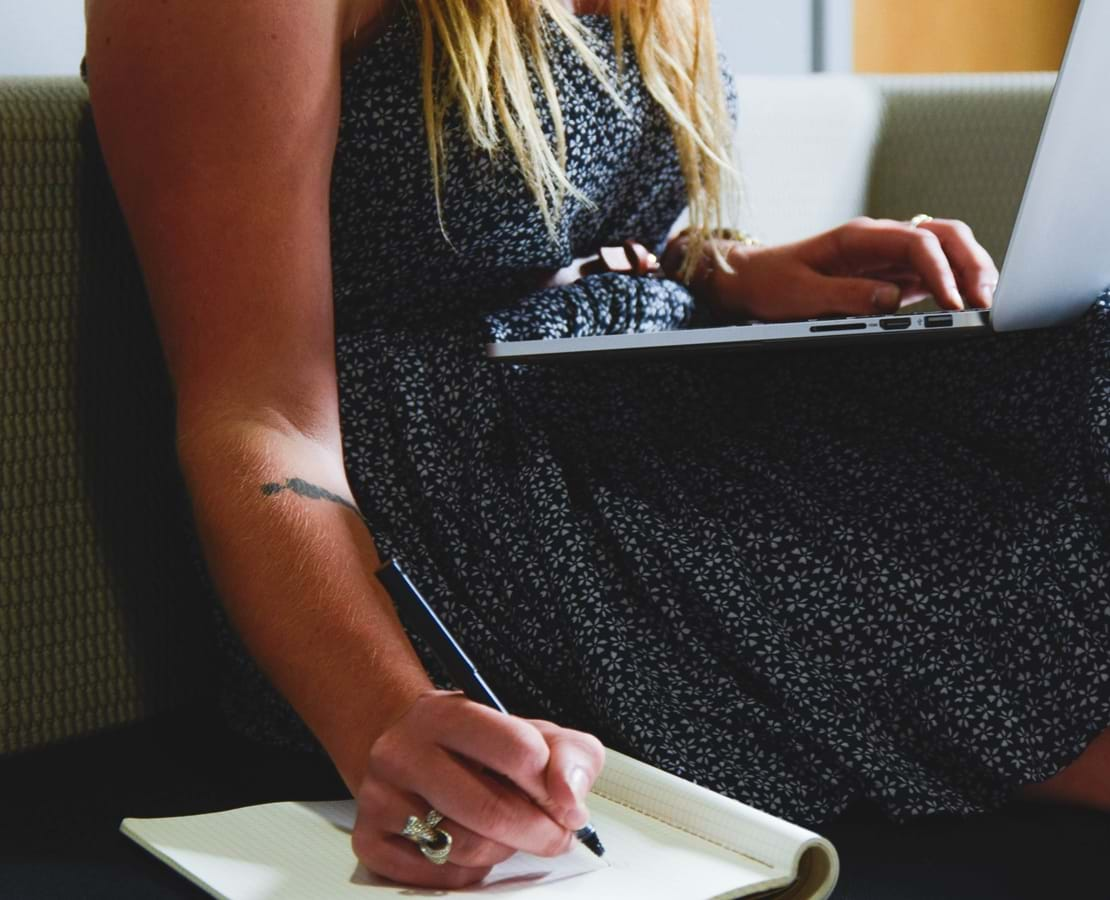 Kvindelig studerende sidder i en sofa med en laptop på skøddet og tager noter i et hæfte der ligger ved siden af hende.