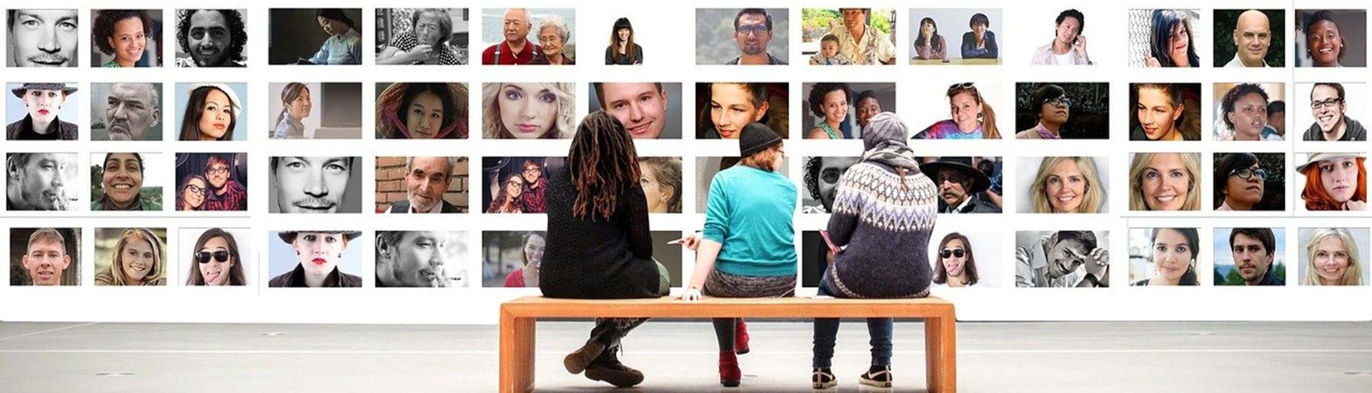 Tre mennesker sidder på en bænk og kigger på en væg fuld af ansigter