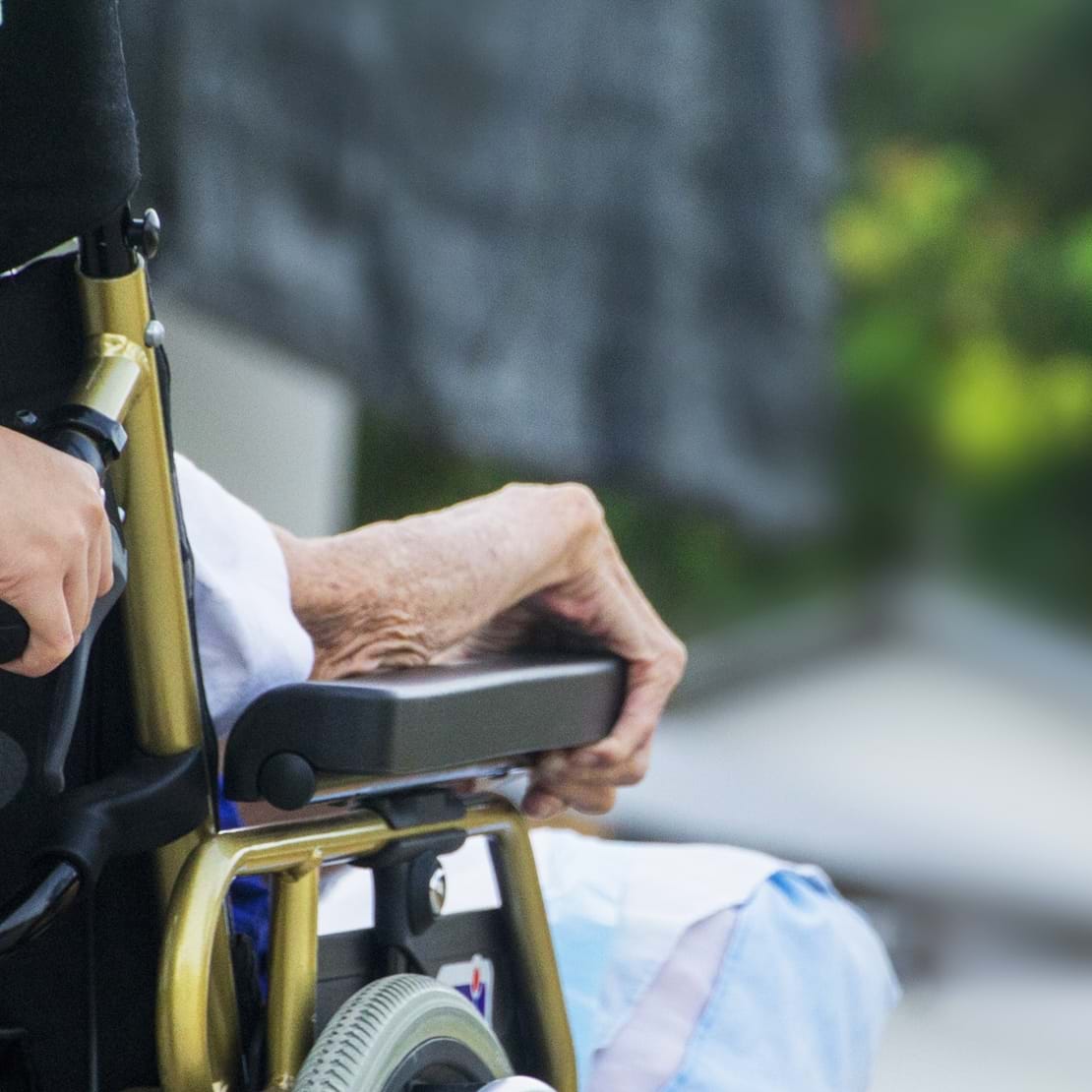 Ældre mand i kørestol bliver skubbet af en SOSU-hjælper hen ad en sti.
